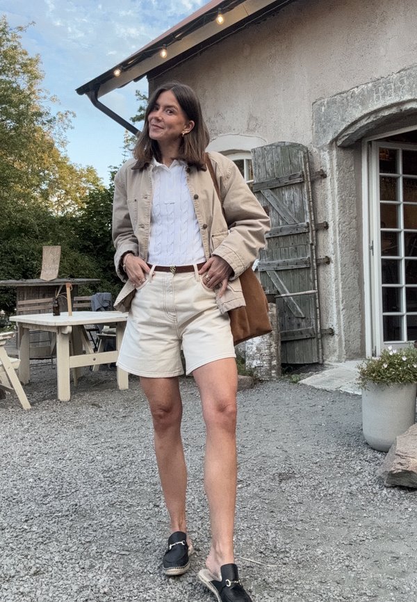 Giacca beige sopra una maglia bianca a treccia, pantaloni shorts crema e scarpe nere senza lacci. Cintura marrone e borsa marrone. Ambientazione all'aperto con ghiaia.
