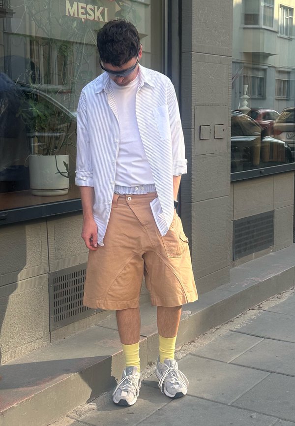 Ung mand iført solbriller, stribet skjorte over hvid T-shirt, beige shorts, gule sokker og sneakers står på en byfortov og kigger ned.