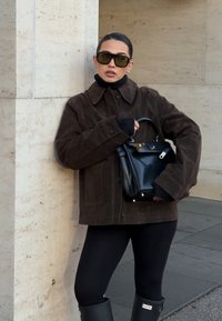 Brun semsket jakke med krage, svart høyhalset genser og svart håndveske. Tilbehør inkluderer store mørke solbriller. Svarte leggings og gummistøvler.