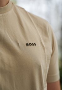 Beige kortärmad T-shirt gjord av mjukt material med rundad halsringning, med en svart "BOSS" logotyp på övre vänstra sidan.