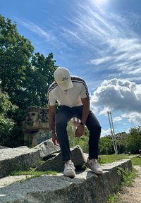 Polo bianco zip-up con strisce grigie, pantaloni scuri e sneakers bianche. Sullo sfondo ci sono vegetazione e una scultura in pietra. Cielo luminoso.