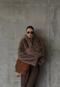 Veste en fausse fourrure marron avec un col montant, portée sur un pantalon marron foncé. Accompagnée d'un sac à épaule en daim marron et de lunettes de soleil rondes.