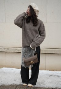 Femme portant un béret beige, un pull marron à texture duveteuse, un pantalon noir avec une superposition en dentelle, et des baskets blanches, tenant un sac à main en cuir marron.
