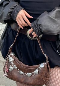 Καφέ δερμάτινη τσάντα με ασημένιες λεπτομέρειες αλυσίδας σε σχήμα καρδιάς, υφή με κείμενο και μια μπρελόκ με γοητεία του Πύργου του Άιφελ. Φορεμένη με μαύρη φούστα.