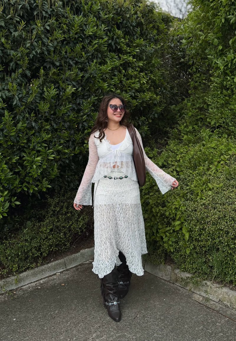 Femme en robe blanche en dentelle, bottes noires et lunettes de soleil souriante, debout sur un chemin entouré de buissons verts denses.