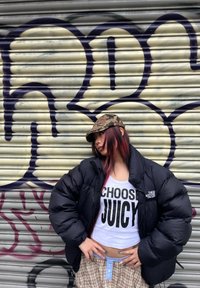 Casaco puffer preto, top curto branco com o texto "ESCOLHE JUICY", calças à quadrados, boné camuflado, cabelo longo com mechas cor-de-rosa, e fundo de graffiti.