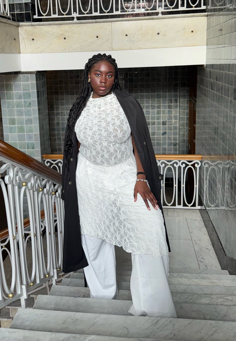 Robe à superposition de dentelle blanche associée à un pantalon blanc ample et un long manteau noir, présentant un motif texturé et une silhouette fluide.