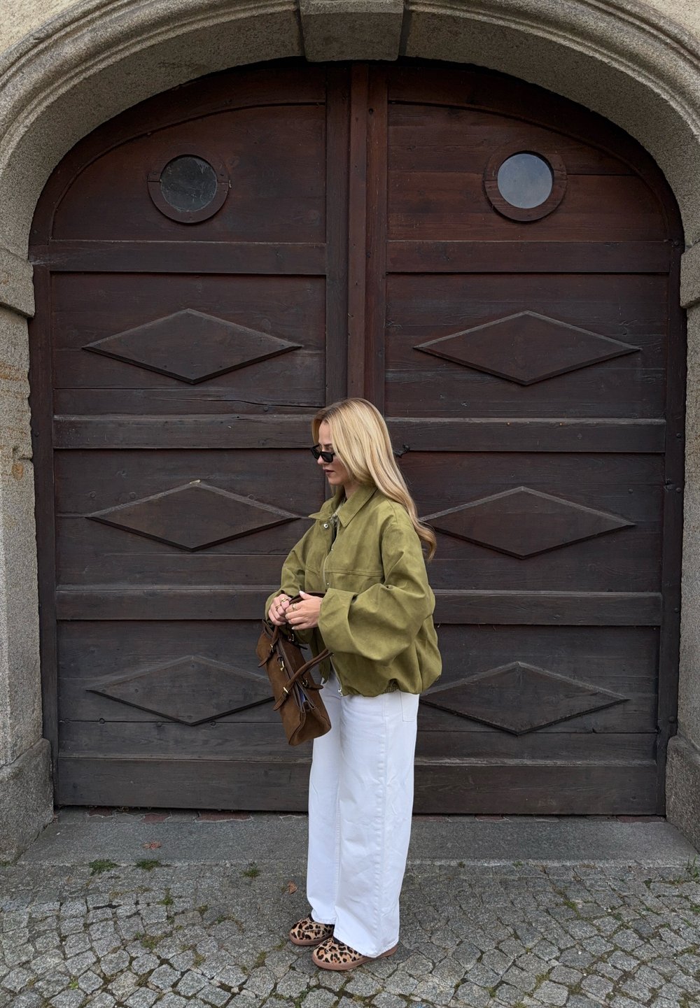 Maslinasto zelena oversize jakna, bijele široke hlače, smeđa torba i sandale s uzorkom leoparda; stoji ispred velikih drvenih vrata.