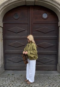 Maslinasto zelena oversize jakna, bijele široke hlače, smeđa torba i sandale s uzorkom leoparda; stoji ispred velikih drvenih vrata.