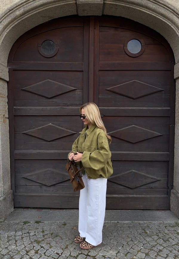 Olivno zelena prevelika jakna, beli hlače širokih hlačnic, rjava torbica in sandali z leopardjim vzorcem; stoji pred velikimi lesenimi vrati.