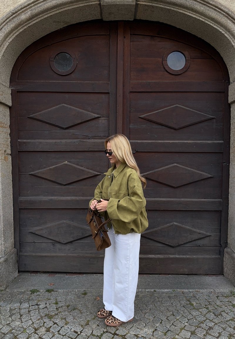 Casaco oversized verde-oliva, calças brancas de perna larga, mala castanha e sandálias com padrão de onça; em pé em frente a grandes portas de madeira.