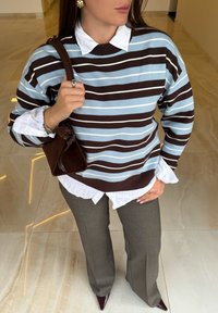 Pull rayé bleu clair et marron, superposé sur une chemise blanche à col. Pantalon évasé gris avec chaussures marron pointues. Sac marron tenu en main.