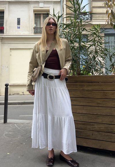 Chaqueta beige clara, top burdeos, falda blanca de capas, cinturón negro con hebilla plateada, zapatos marrones. El fondo presenta vegetación y un entorno urbano.