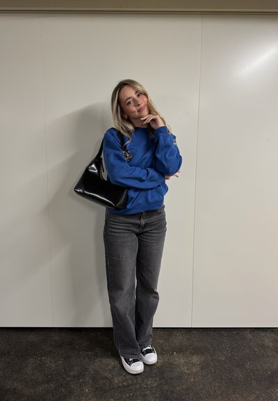 Sudadera azul, jeans grises, bolso negro y zapatillas blanco y negro. De pie contra una pared lisa, con un brazo doblado y la barbilla apoyada en la mano.