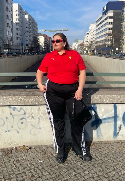 Camisa polo roja con el logo de Ferrari, pantalones de chándal negros con rayas blancas, bolso negro y gafas de sol rojas. Fondo urbano con graffiti.