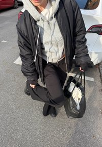 Halli hallis hulka ja musta jope kandja, kes hoiab musta nahast kotti, seisab asfaldil pargitud valgete ja punaste autode lähedal.