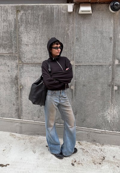 Persona con sudadera negra con capucha, gafas de sol y vaqueros de pierna ancha, de pie con los brazos cruzados frente a una pared de hormigón, lleva una bolsa negra grande.
