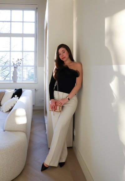 Top negro de un solo hombro con detalle de volantes, pantalones blancos de talle alto, tacones negros puntiagudos y un bolso pequeño beige con cadena.