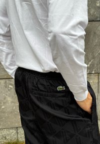 Calças de desporto pretas com um padrão geométrico, cintura elástica e bolsos laterais. Logótipo verde da Lacoste no bolso traseiro. Acompanhadas por uma camisa branca de mangas longas.