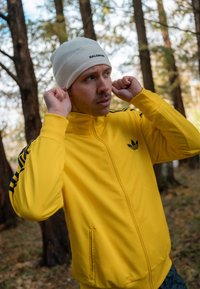 Żółta sportowa kurtka z czarnymi akcentami, pełnym zamkiem błyskawicznym i stójką. Biały czapka beanie z logo. Naturalne tło na świeżym powietrzu.