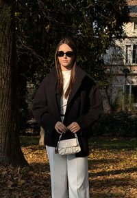 Palton negru supradimensionat peste un top alb, asortat cu pantaloni albi. Accesoriile includ o geantă mică albă și ochelari de soare închiși la culoare.