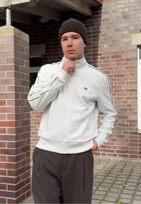 Homem a usar uma jaqueta branca com fecho, um boné castanho e calças escuras, de pé ao ar livre perto de colunas e uma parede de tijolos, com a mão perto do queixo.