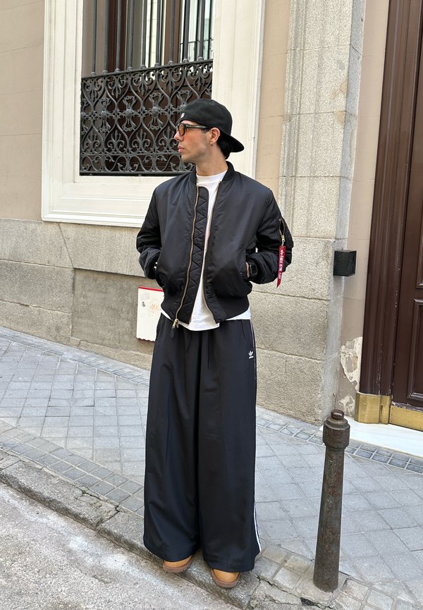 Casaco bomber preto com um acabamento texturizado, t-shirt branca, calças de treino pretas largas com riscas e calçado castanho. De pé contra uma parede de pedra.