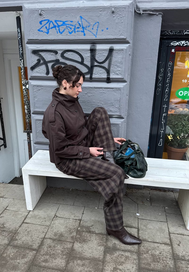 Persona con giacca marrone e pantaloni a quadri seduta su una panchina bianca, che si sistema lo stivale, con una borsa verde e un muro grigio coperto di graffiti sullo sfondo.