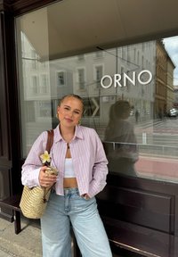 Lichtroze gestreepte overhemd met knopen over een witte crop top, gecombineerd met hoog-gewaste lichtblauwe jeans. Houdt een geweven tas met een bloemenaccent vast.