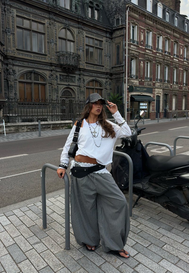 Femme en pantalon ample à carreaux, top court blanc et casquette, appuyée sur une rampe de rue à côté d'un scooter garé avec des bâtiments historiques ornés en arrière-plan.
