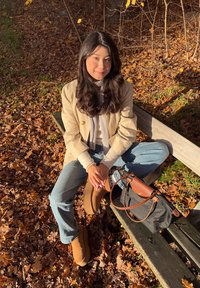 Nuori nainen beige takki, huivi ja saappaat päällään istuu penkillä syksyn lehtien keskellä, vieressään musta ja ruskea käsilaukku.