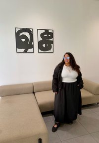 Vrouw met lang krullend haar, die een bril draagt, in een witte top, zwarte jas en rok, staat bij een beige sectionele bank in een minimalistische kamer met abstracte kunst.