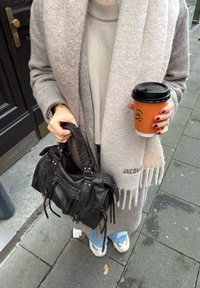 Bolso negro con textura y asa trenzada, bufanda de lana gris, suéter de punto beige y una taza de café con funda naranja, colocados sobre el pavimento.
