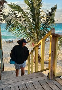 Mann in schwarzem Kapuzenpullover und Shorts, der eine Holztreppe vom Sandstrand hinaufgeht, im Hintergrund eine Palme und türkisfarbenes Meer.