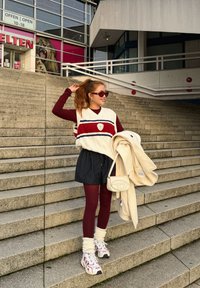 Persoană pe scări purtând un vest tricotat crem cu dungi roșii și albastre, mâneci lungi bordo, pantaloni scurți negri cu dungi, colanți bordo și adidași cu talpă groasă.
