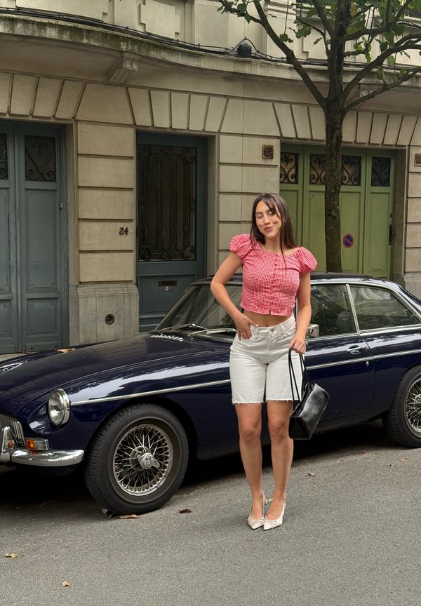 Donna con top a quadri rossi e pantaloncini bianchi in piedi sulla strada accanto a un'auto d'epoca blu scuro e un albero davanti a edifici residenziali.