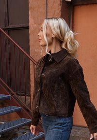 Pruun seemisnahast jakk kraega kaelusega, nööpidega kinnitatud ja küljetaskutega; kantud tumeda särkriide peal ning koos teksapükstega.