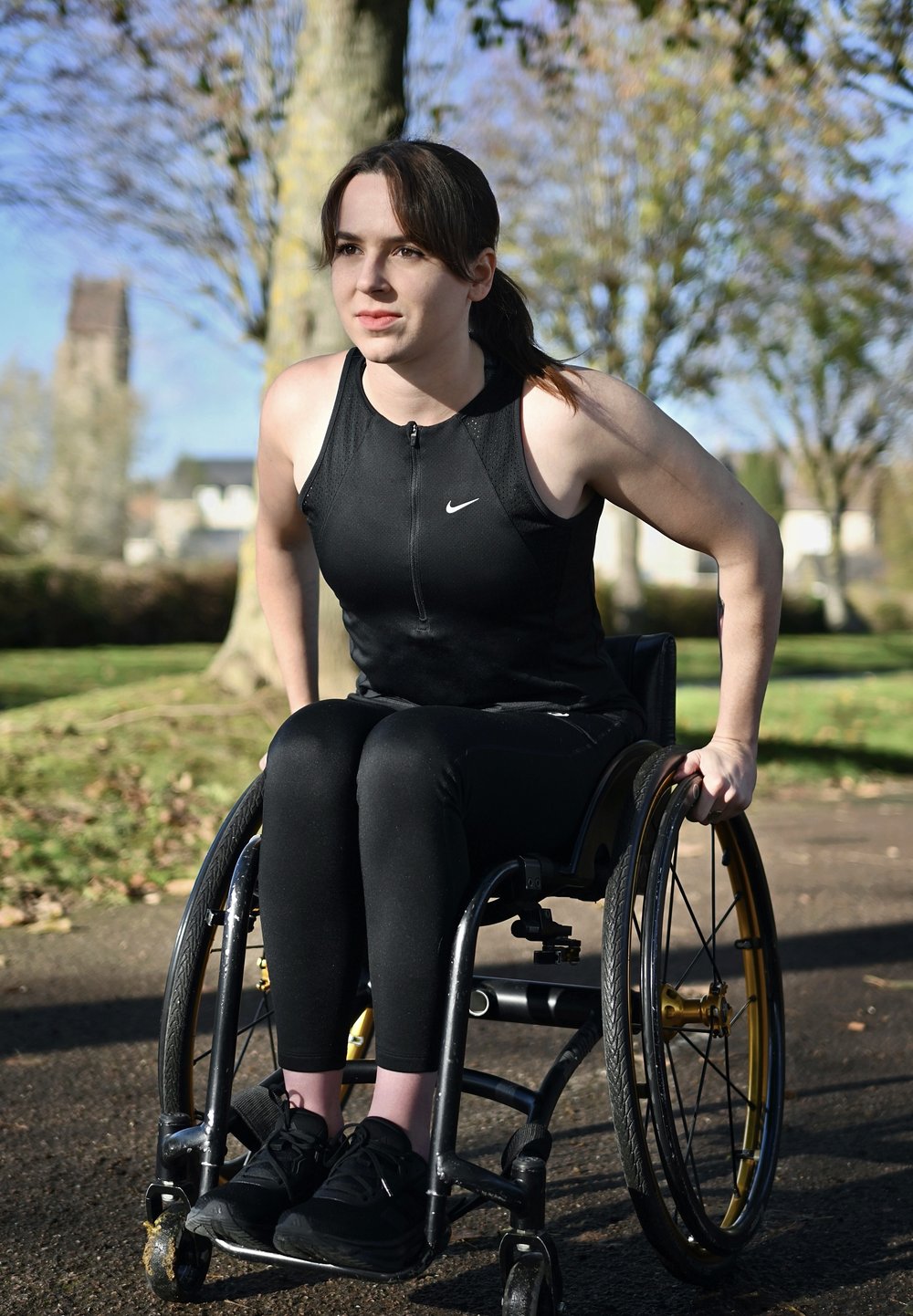 Débardeur de sport noir avec fermeture éclair, leggings noirs et baskets noires. Personne utilisant un fauteuil roulant avec des roues noires et dorées, tenant les poignées.