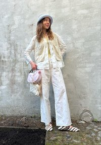 White embroidered blouse paired with a cream knitted vest, wide-fitting white trousers, striped brown and white shoes, and a pastel crochet hat. Two bags are being held.
