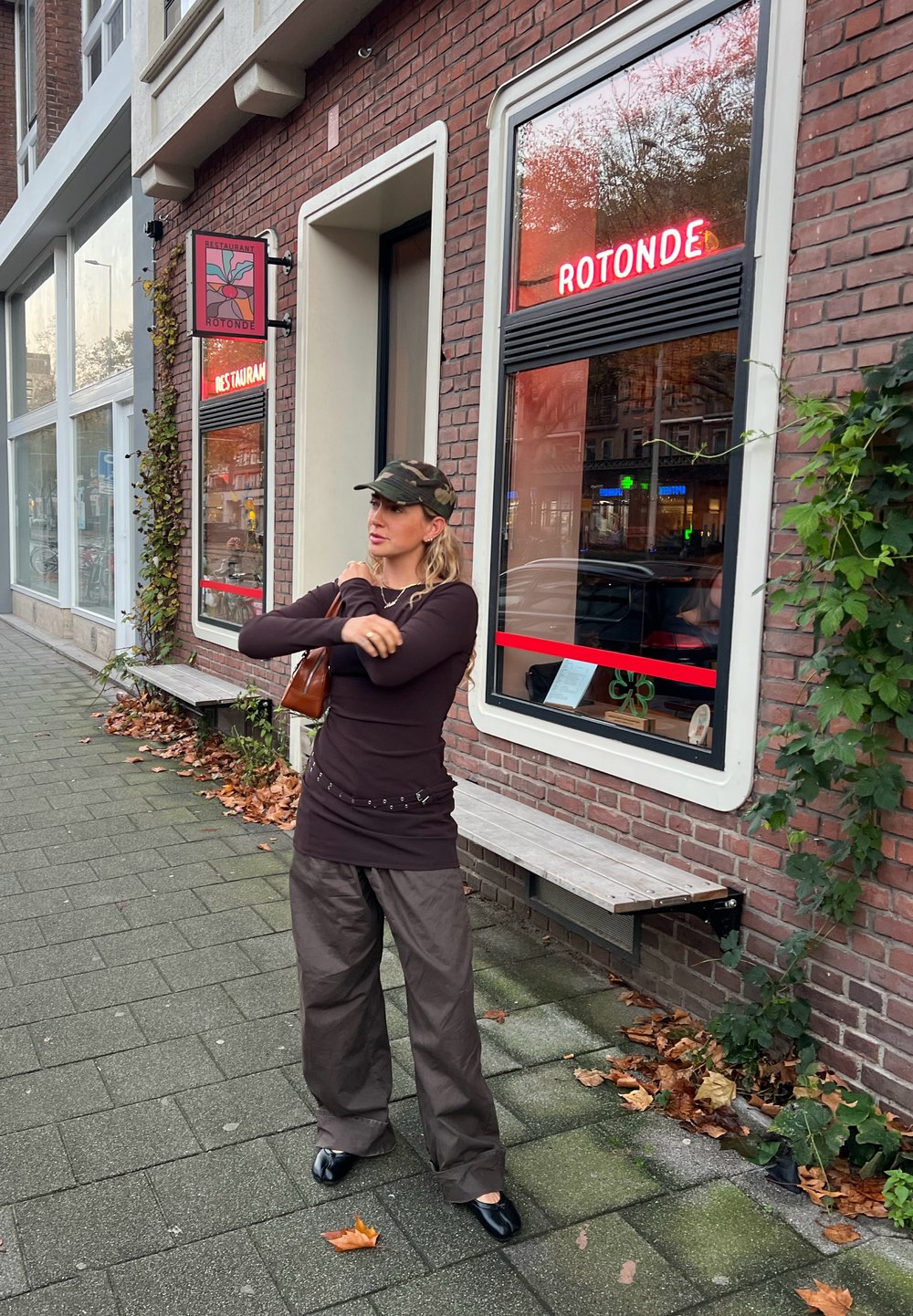 Femme portant une casquette camouflage et une tenue sombre, debout sur le trottoir devant un restaurant avec un néon rouge indiquant "ROTONDE", feuilles d'automne au sol.