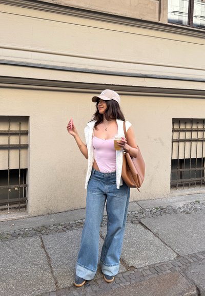 Chaqueta sin mangas blanca sobre una blusa rosa, jeans de pierna ancha de color azul claro, bolso marrón y gorra beige, sosteniendo una bebida y un teléfono.