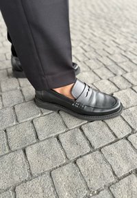 Mustat nahka loaferit, joissa on sileä pinta, varustettu elastisilla nauhoilla ja pyöreällä kärjellä. Yhdistetty mustien housujen kanssa tekstuuripintaiseen kivipintaan.