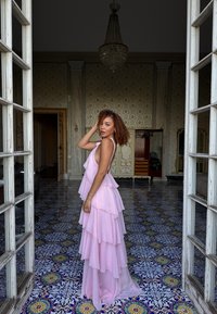 Vestido rosa de capas con volantes, hecho de tela ligera. El vestido cuenta con un escote halter y una silueta fluida, situado sobre un suelo de azulejos ornamentados.