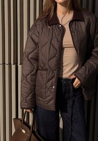 Brun quiltet jakke med mørk krage, knapper og lomme foran, kombinert med en beige topp og mørkeblå vide bukser, mens hun holder en brun veske.