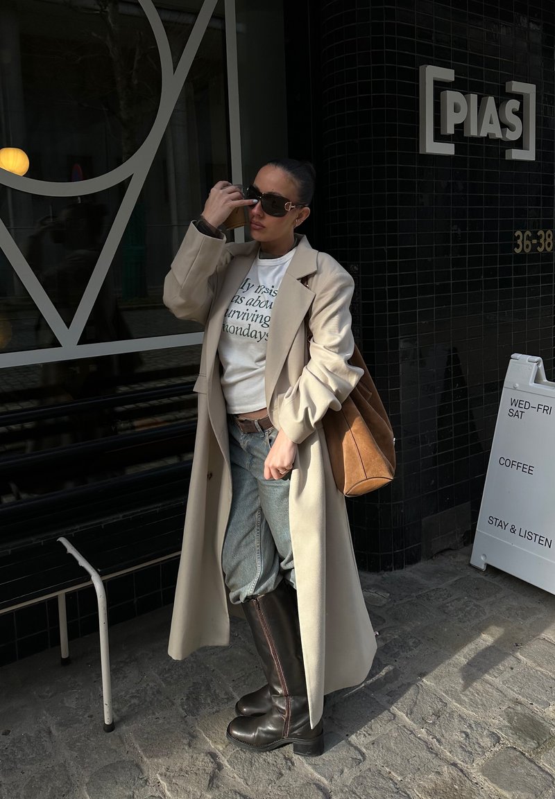 Kvinne som justerer solbriller, iført lang beige frakk, hvit skjorte med trykk, jeans og knelange svarte støvler, stående utenfor en kafé med skilt.