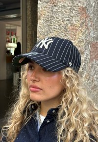 Ung kvinna med långt lockigt blont hår som bär en svart keps med vita ränder och en vit logotyp, mörk jacka och kedjehalsband, stående mot en stenvägg.