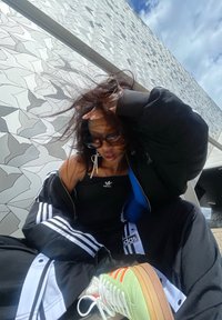 Pantalons de jogging noirs Adidas avec des rayures blanches, un débardeur noir et des baskets rétro colorées ; en arrière-plan, un mur à motifs géométriques.