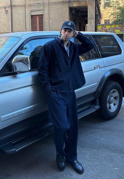 Chaqueta de mezclilla oscura y pantalones, gorra negra, camisa de cuadros blanca debajo, zapatos de cuero negros, de pie al lado de un SUV plateado.