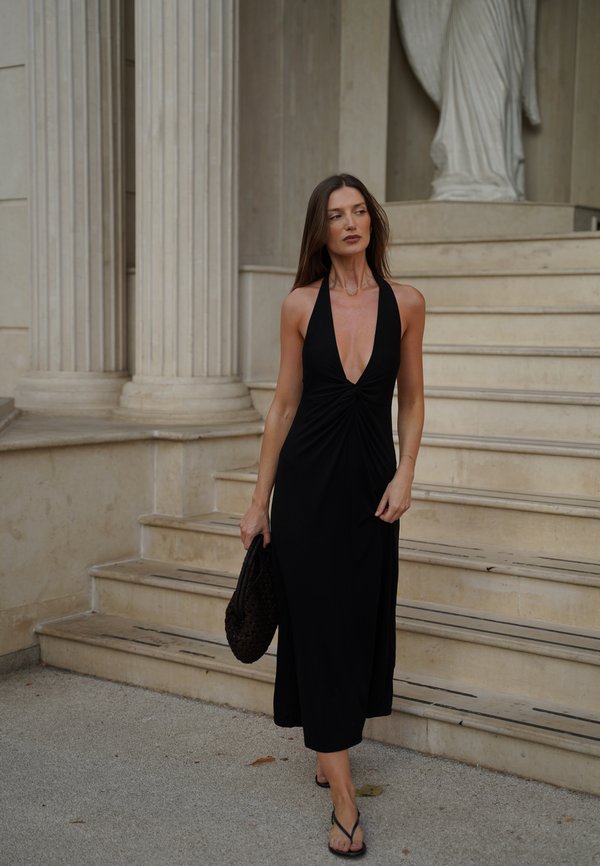 Frau in einem schwarzen Neckholderkleid, die eine schwarze Tasche hält, läuft eine Steintreppe nahe großer klassischer Säulen und einer Statue hinunter.