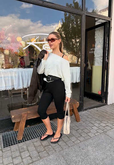 Joven mujer con gafas de sol sostiene una chaqueta de cuero sobre el hombro y una bolsa, parada en una calle adoquinada frente a una cafetería con una ventana grande.